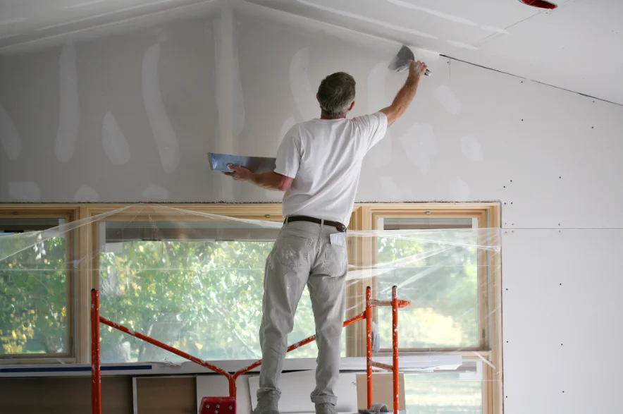 Drywall finisher taping a vaulted ceiling with the windows sealed off by plastic sheeting